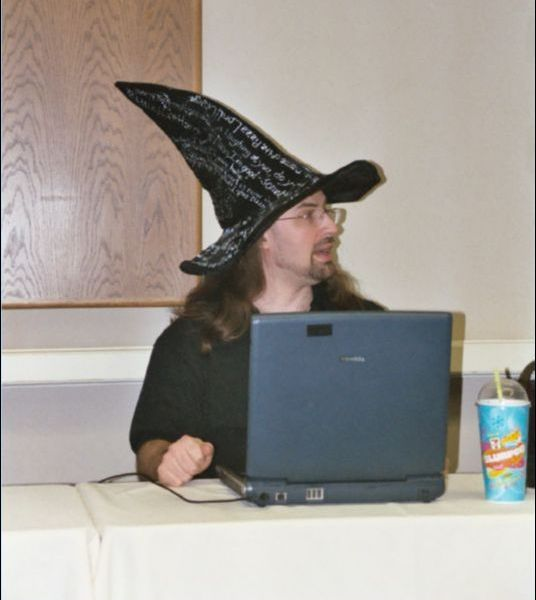 Jim Butcher wearing a black wizard hat with Dresden Files quotes painted on it in silver paint; he's sitting at a table with a laptop open in front of him