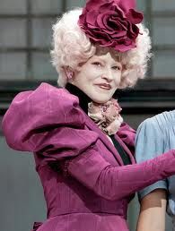 shot of Effie from film adaption of The Hunger Games in purple dress with white hair and giant crazy flower atop her head wtf is that