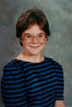 image of me as a 6th-grader, with short hair, big glasses, and a blue-and-black striped dress