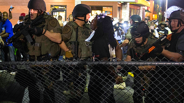 image of four white male police officers in riot gear holding assault rifles; two of them are pointing their weapons toward protesters who surround them, while one handcuffs a black man against a chain-link fence and the other looks at protesters behind them 