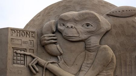 image of a sand sculpture of E.T. 'phoning home' by pressing a long finger against a telephone keypad