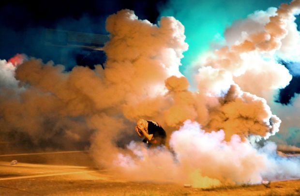 image of a protestor crouching down in the middle of a street, as clouds of tear gas engulf hir