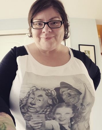 image of me standing in my entryway wearing a 3/4 sleeve ringer shirt with a photo of the characters from The Wizard of Oz posing for a selfie while Dorothy holds out a cellphone