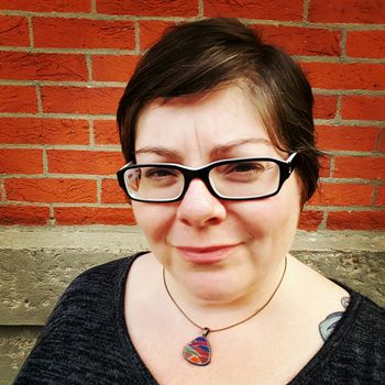 image of me standing in front of a brick wall outdoors, wearing a grey scoop-neck top and a necklace with a triangular bauble of colored glass