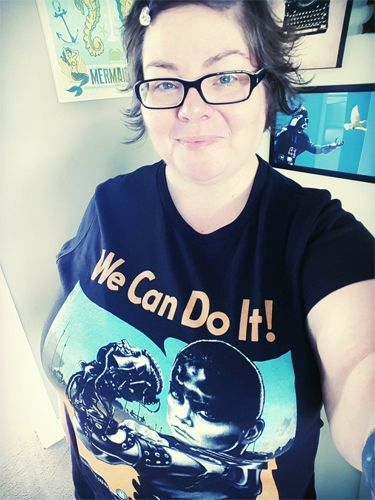 image of me standing in my living room wearing a t-shirt featuring Furiosa from Mad Max: Fury Road lifting her arm in the fashion of Rosie the Riveter and the text We Can Do It!