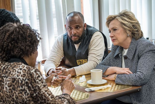 image of Hillary Clinton sitting at a table beside a black man, across from two black women; one of the women is talking, and Hillary and the man are looking at her and listening intently
