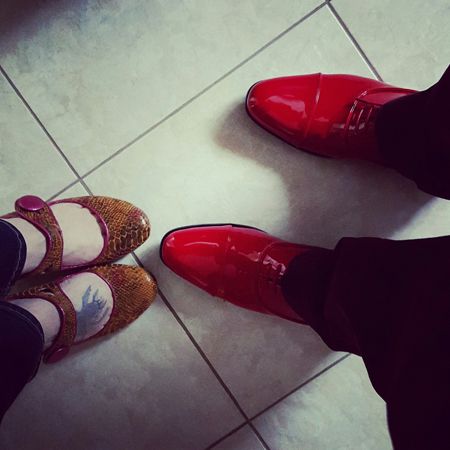 image of two sets of feet: a man's feet wearing shiny red shoes and a woman's feet wearing pink textured MaryJanes
