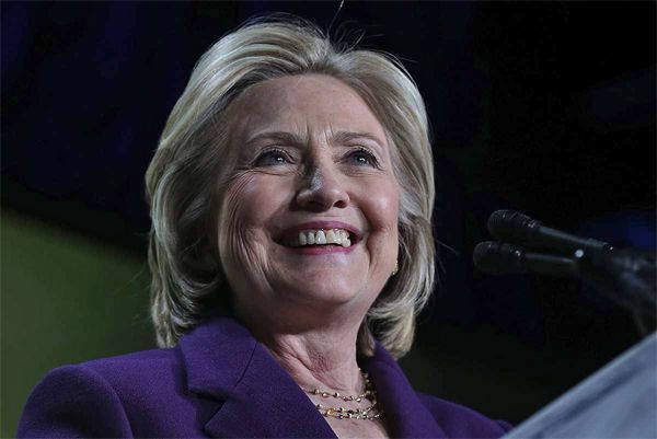 image of Hillary Clinton standing at a podium smiling, wearing a purple jacket