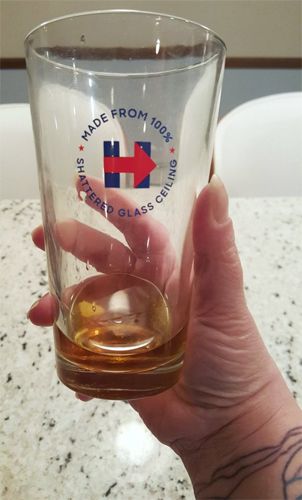 image of my hand holding a glass with a shot of whiskey in it; on the glass is Hillary Clinton's logo accompanied by text reading: 'Made from 100% shattered glass ceiling.'