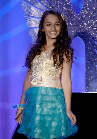 image of Jazz Jennings, a brown-skinned teenage girl with long brown hair, wearing a dress with a gold patterned top and turquoise ruffled skirt