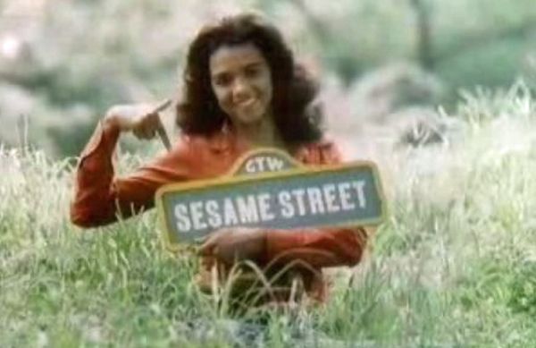 1970's image of Sonia Manzano, playing Maria, standing in a grassy field holding a Sesame Street sign