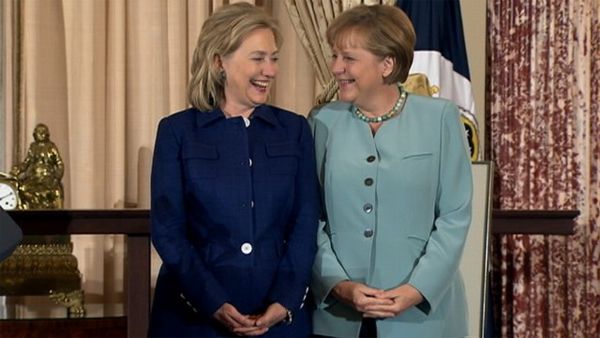 image of Hillary Clinton standing beside German Chancellor Angela Merkel, an in-betweenie white woman; they are standing in the same position, their hands clasped in front of them, looking at each other and smiling