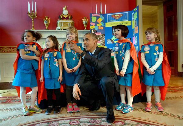 image of President Obama kneeling and pointing at something out of frame, surrounded by five little girls of various ethnicities, who are wearing blue uniforms and red capes, all looking in the direction he is pointing