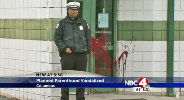 image of a black male police (or security) officer standing outside the front door of the building, which has been splashed with red paint