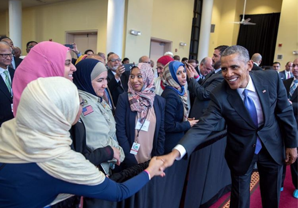 image of President Obama smiling broadly while shaking hands with a Muslim woman wearing a headscarf, who is standing beside three other Muslim women in headscarves, behind a barrier separating the President from a crowd of people at a mosque