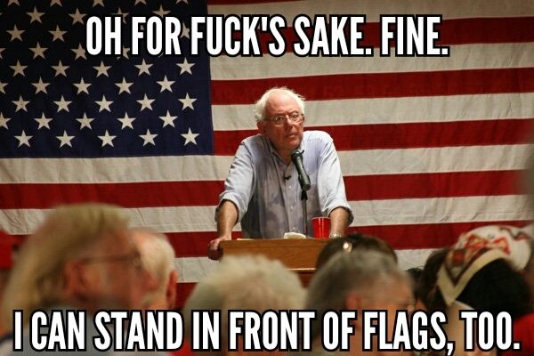image of Bernie Sanders, standing at a podium during a speaking event, in front of a giant US flag, with a perturbed look on his face, to which I've added text reading: 'Oh for fuck's sake. Fine. I can stand in front of flags, too.'