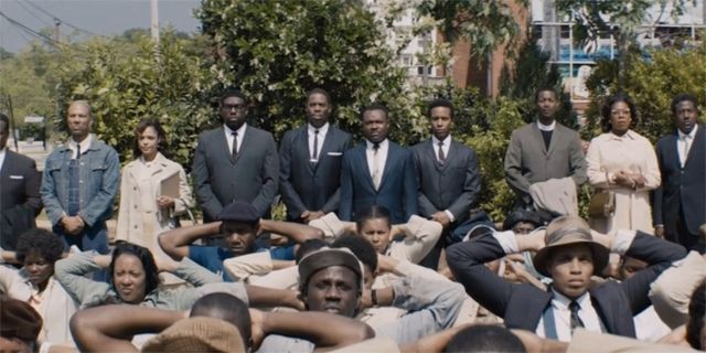 screen cap from the film Selma, showing Martin Luther King Jr. (David Oyelowo) and other members of the SCLC standing at the back of a group of black people, who are kneeling down in protest, with their hands raised and behind their heads, in front of the Selma courthouse
