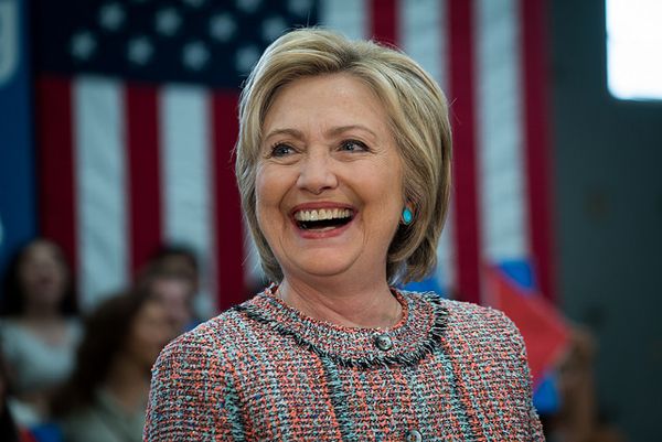 image of Hillary Clinton at a campaign event, surrounded by people with a US flag in the background; she is smiling broadly