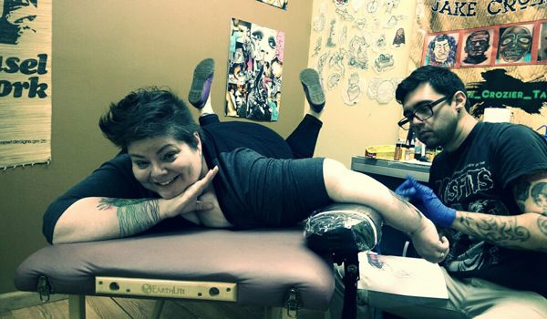 image of me lying on Jake's table, making a silly face with my feet in the air, while Jake tattoos me