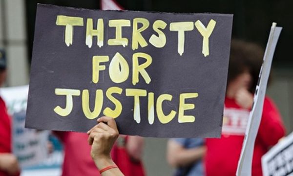 image of a protestor in Detroit holding up a handwritten sign reading: THIRSTY FOR JUSTICE.