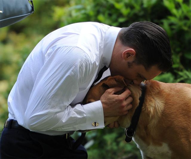 image of actor Tom Hardy, dressed in a period button-down, tie, and slacks, leaning over to kiss the top of his golden mutt's head