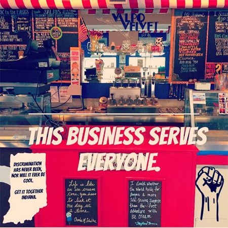 image of Valpo Velvet Ice Cream's storefront, with a sign shaped like Indiana reading 'Discrimination has never been, nor will it ever be, cool. Get it together, Indiana.' and large letters on the window reading 'THIS BUSINESS SERVES EVERYONE.'