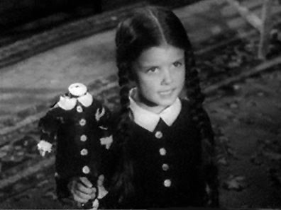 screen shot of Wednesday Addams from the old television show, holding up a doll which is missing its head