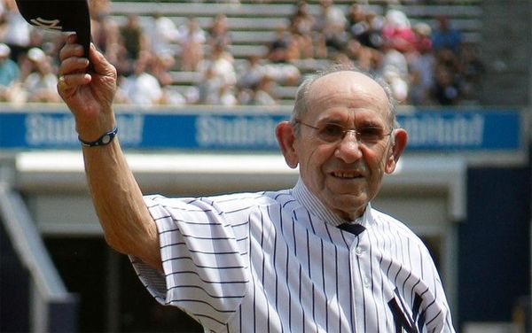 image of Yogi Berra standing in a stadium, wearing a Yankees uniform, and waving his cap while smiling at the crowd