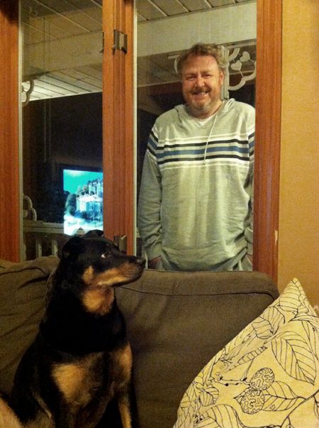 image of Iain standing outside the window, smiling; just below, inside, Zelda sits on the loveseat looking dubious