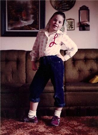 image of me at age 9, standing in my living room in a sassy pose, with my hand on my hip, my hair tied up in a scarf, my jeans rolled up, and sporting a blouse with a cursive L on it