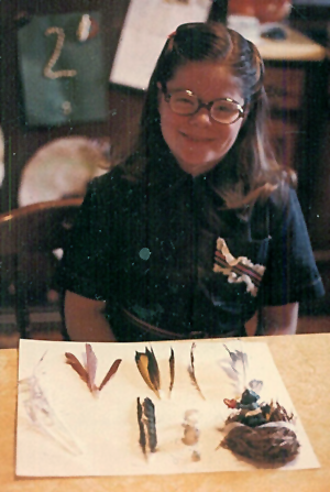 image of me as a little girl, age 9, wearing a denim jumpsuit and round glasses, with an array of stuff collected from the outdoors spread out in front of me on our kitchen table