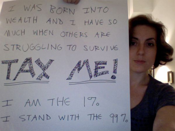 image of white woman holding a sign reading 'I was born into wealth and I have so much when others are struggling to survive.  TAX ME!  I am the 1%.  I stand with the 99%.'