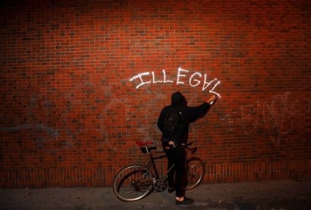 a person in dark clothes, standing with a bicycle, sprays 'illegal,' with upside-down A, on a brick wall in Oakland