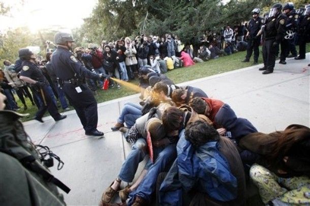 a police officer sprays a group of protesters, sitting peacefully on the ground, with pepper spray