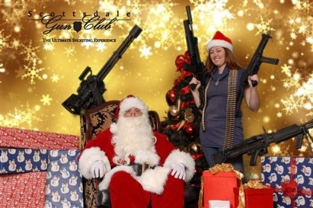 a picture of a white woman in a Santa cap posing with a Santa Claus while holding huge guns