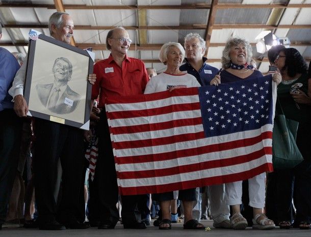a few older white people, holding a US flag (backwards) and a framed portrait of Ronald Reagan, smile excitedly while waiting for Newt Gingrich to come onstage at a campaign event in Florida