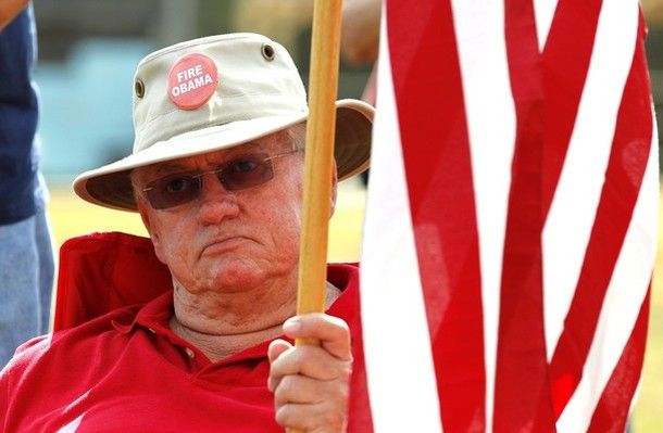 image of older white man sitting in the sun looking grumpy, holding a US flag and wearing a pin on his hat that says 'Fire Obama'