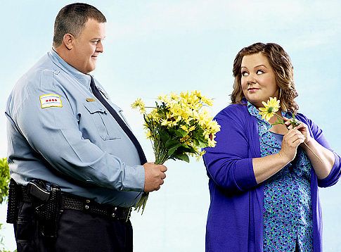 a promotional image for the television show 'Mike and Molly'; Mike is handing Molly a bouquet of flowers