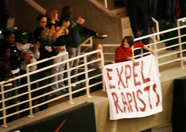 two women in the bleachers at a game holding up a sign reading EXPEL RAPISTS