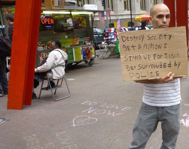 image of an Occupy Wall Street protester holding a sign reading 'Destroy society, get million$. Stand up for justice, get surrounded by police.'