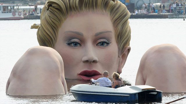image of a giant statue of a white woman comprised of head and knees sticking above water in a lake, with a boat of gawkers floating between her knees