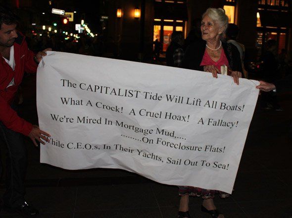image of elderly female Occupy Wall Street protester holding a sign reading 'The CAPITALIST tide will lift all boats! / What a crock! A cruel hoax! A fallacy! / We're mired in mortgage mud...on foreclosure flats! / While CEOs, in their yachts, sail out to sea!'