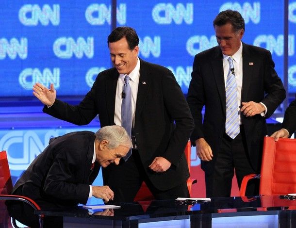 Ron Paul leans forward as Mitt Romney and Rick Santorum walk behind him; Santorum looks like he's about to clap Paul on the back