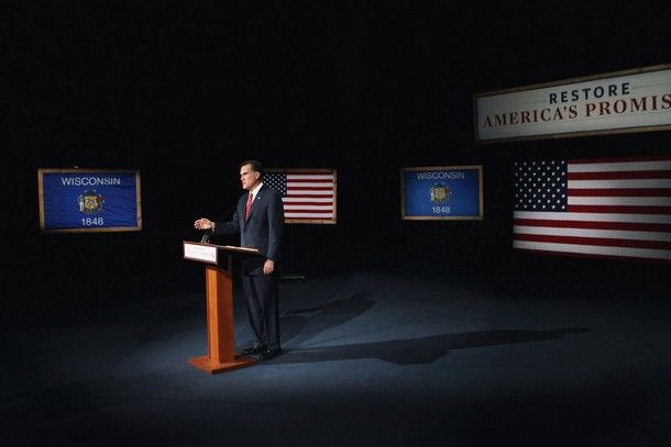 image of Mitt Romney standing at a podium on a dark stage, with two US flags, two Wisconsin state flags, and a banner reading 'Restore America's Promise' hanging randomly around the sparse walls