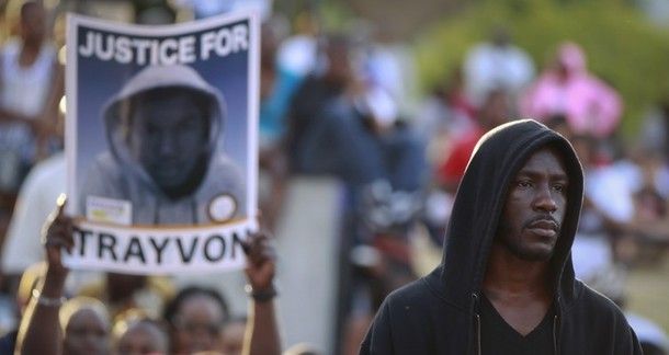 a black man wearing a hoodie stands next to a fellow protestor at a demonstration holding up a sign reading 'Justice for Trayvon' with an image of Trayvon Martin in a hoodie