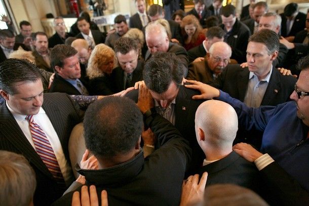 image of Rick Santorum with his head bowed, standing in a circle of people who all have their hands laid on him, their heads bowed, praying