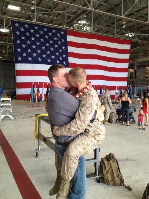 image of two male Marines kissing after one of them, in fatigues, has just returned home from a deployment; in the background is a huge US flag.