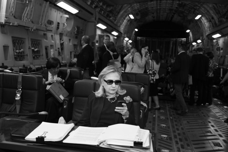image of Hillary Clinton wearing sunglasses and working while in transit, papers in front of her, her phone in her hand