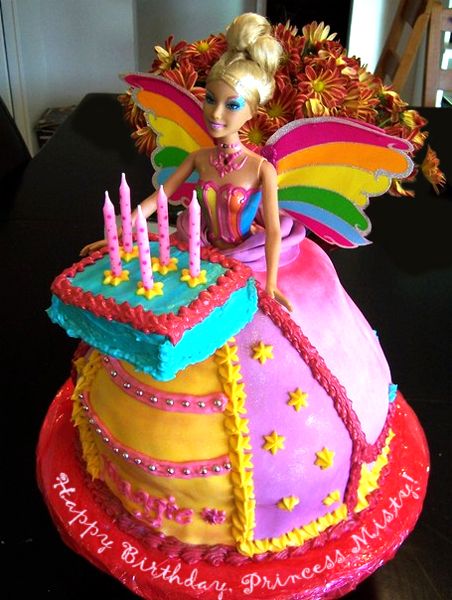 a Barbie cake in which the Barbie is wearing a princess gown, has rainbow wings, and is holding out a birthday cake with candles, labeled 'Happy Birthday, Princess Misty!'
