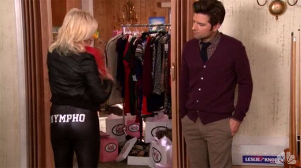 Leslie tries on tight pants reading 'nympho' across the butt and tries to check it out over her shoulder, while Ben looks on curiously